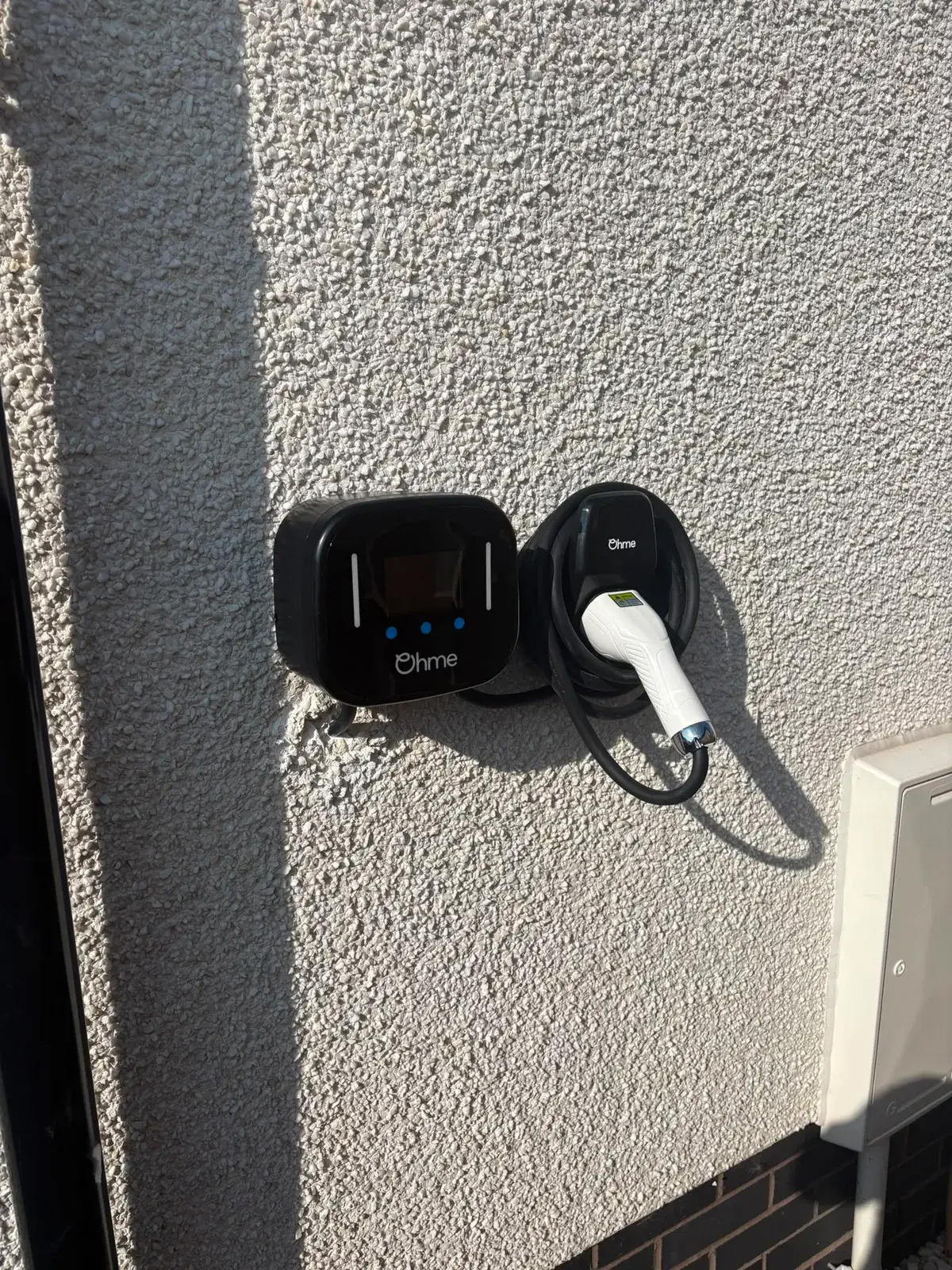 A wall-mounted charge point on a rendered wall with its connector resting in the holster beside it