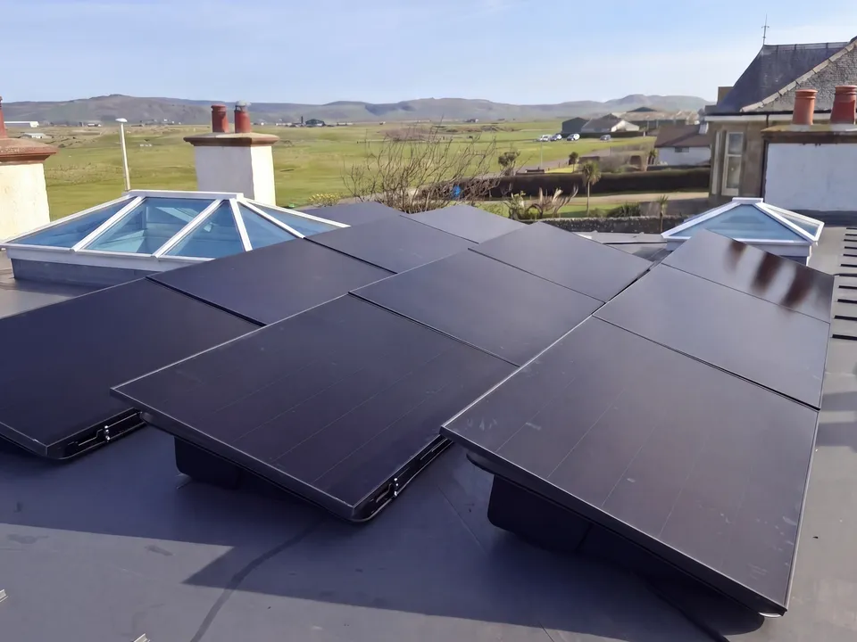 Solar panels on ballasted flat roof mounts beside rooflights, with hills and open fields behind