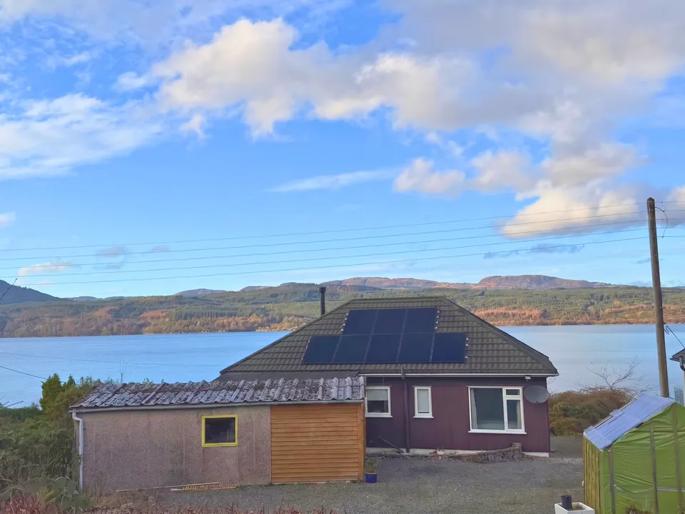 Solar panels on a bungalow roof above a loch, with forested hills across the water